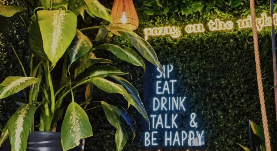Pared de follaje con un letrero de neón que dice Sip, eat, drink, talk and be happy, junto a plantas grandes.