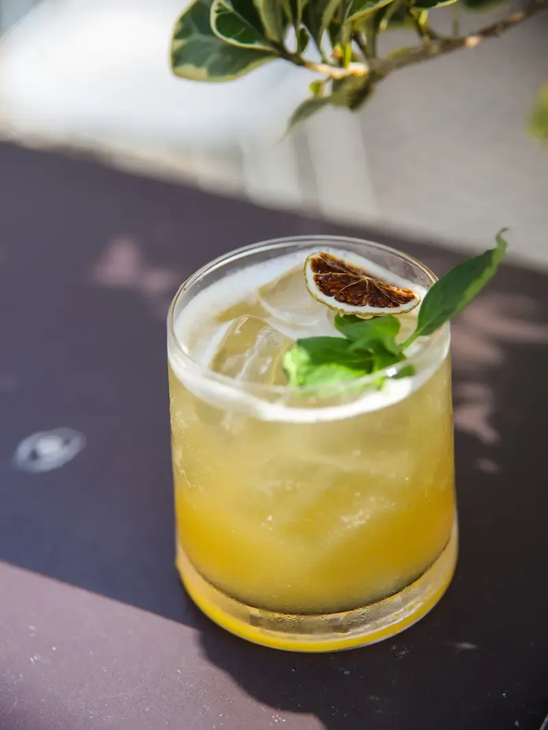 Cóctel dorado con espuma, una rodaja de lima seca y hojas de albahaca.