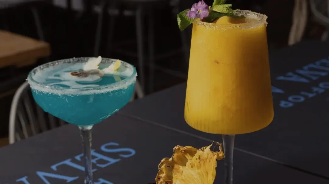 Dos cócteles, uno azul con flores y otro color mango, sobre una mesa con el logo de Selva Azul.