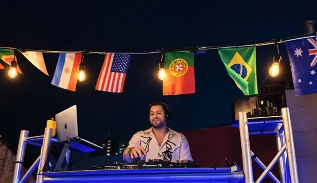 DJ en la cabina del rooftop de noche, bajo banderas y luces.